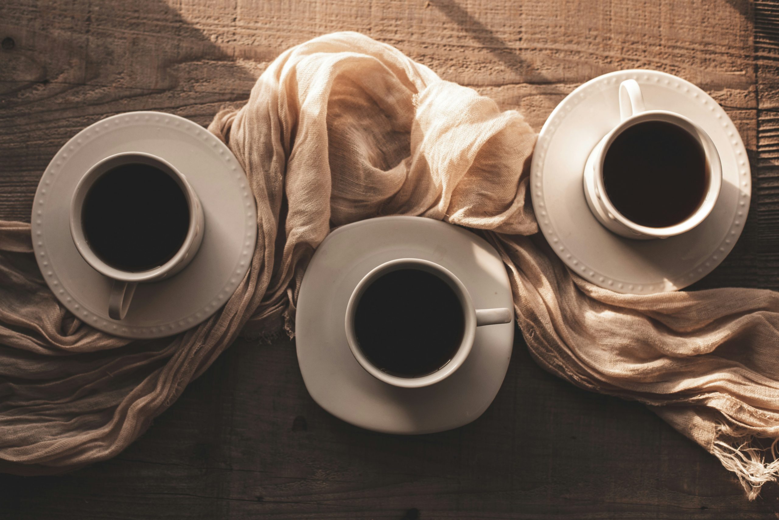 Ansicht in drei gefüllte Kaffeetassen auf einem Tisch. Dazwischen ein Tuch drapiert.