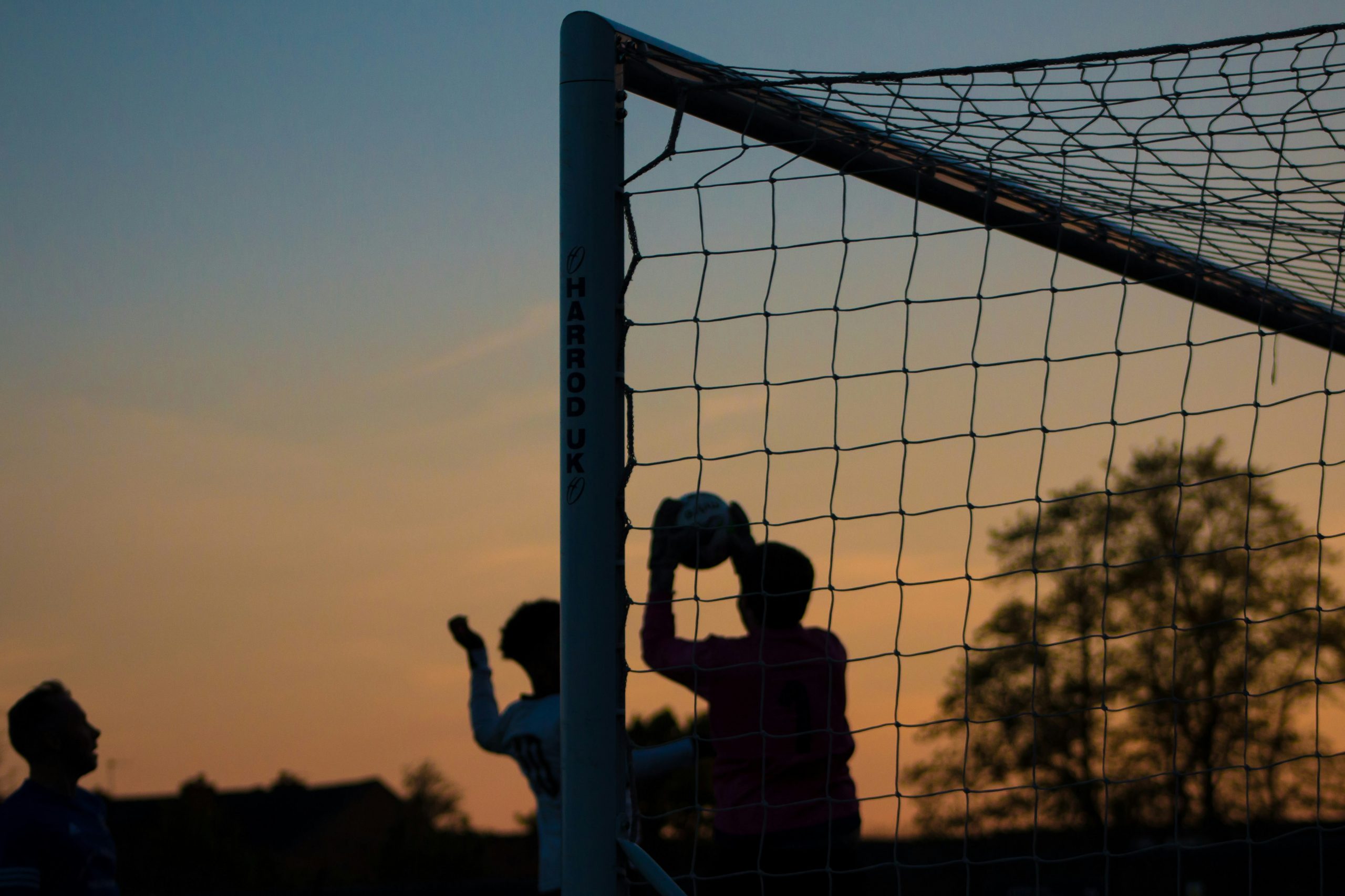Ein Torwart fängt beim Fußballspielen im Sonnenuntergang den Ball, während zwei weitere Spieler vor dem Tor stehen.